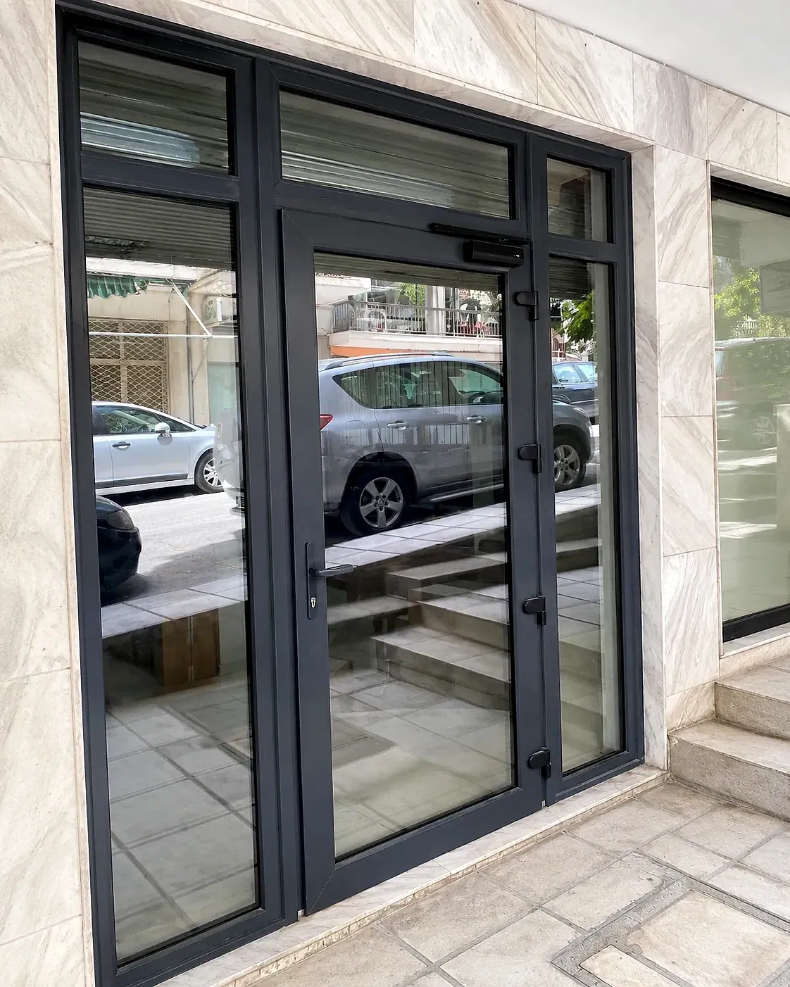Ανθρακί υαλόπορτα αλουμινίου με πλαϊνά σταθερά και φεγγίτες, σε είσοδο κτιρίου.